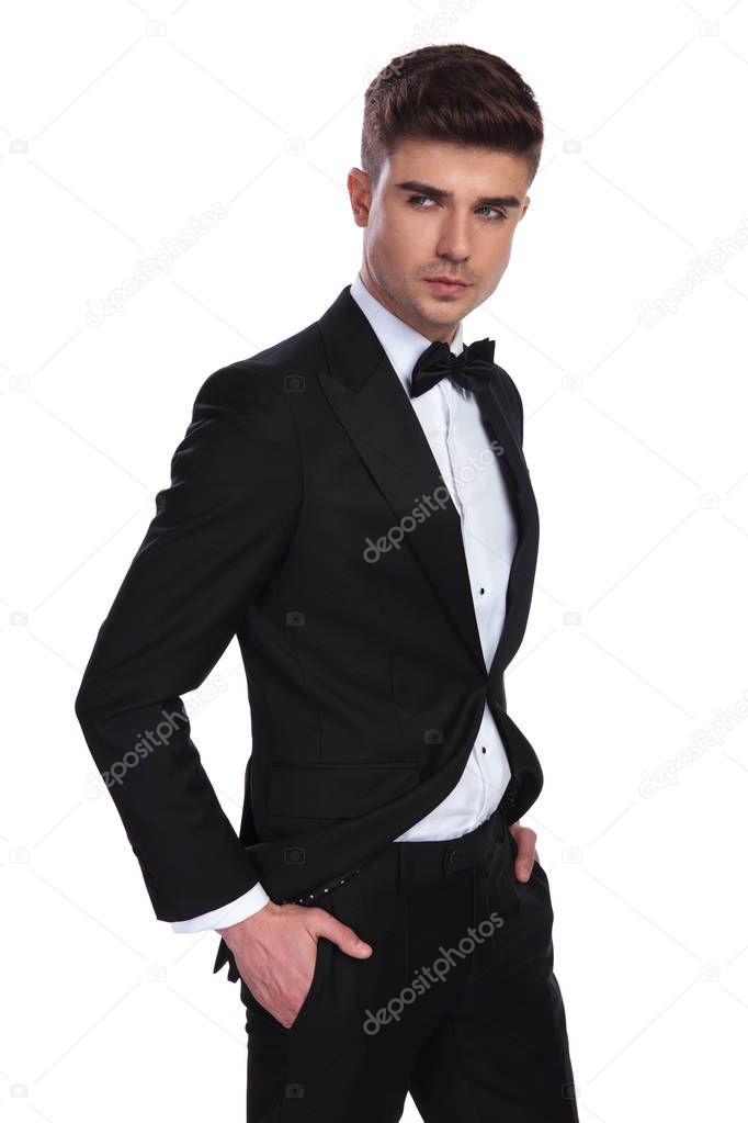 portrait of relaxed classy man wearing a black tuxedo looking to side while standing on white background