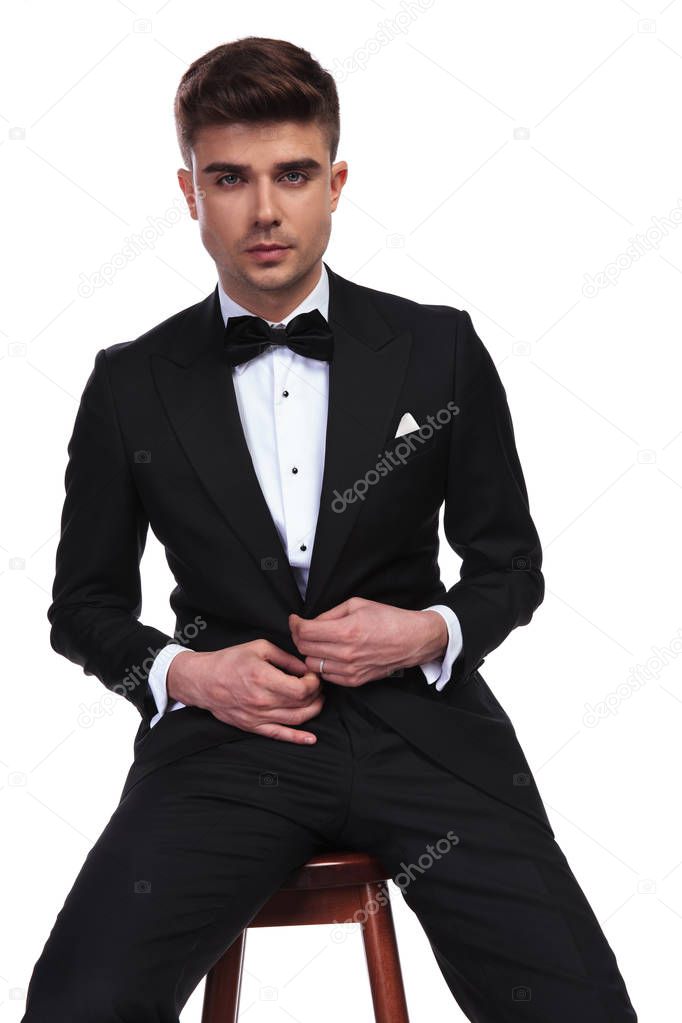 portrait of attractive seated groom buttoning his black suit on white background