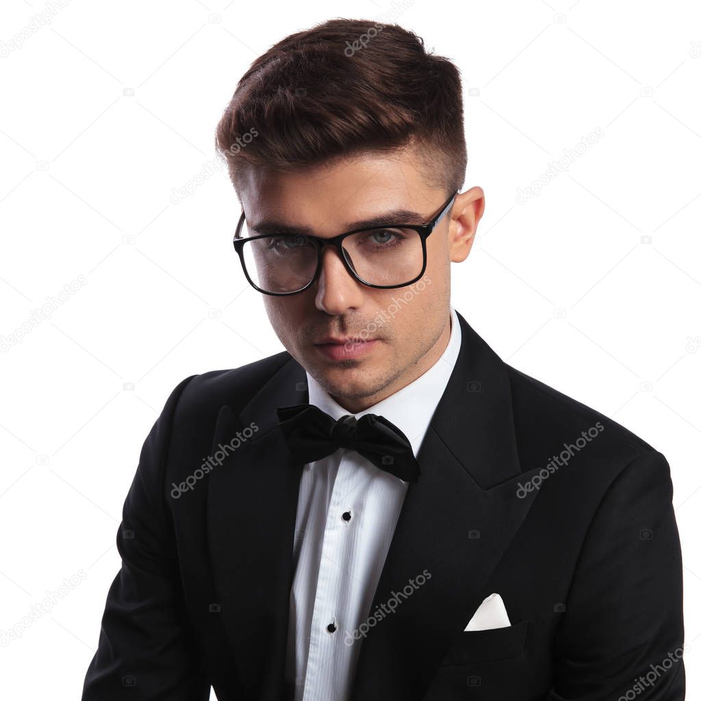 portrait of smart young man wearing black tuxedo and glasses while standing on white background
