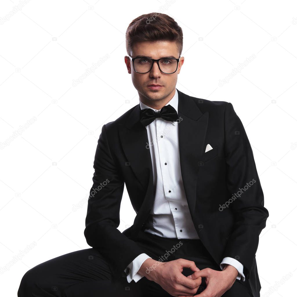 portrait of groom wearing glasses and black tuxedo sitting on white background