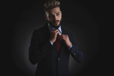 portrait of handsome businessman arranging red tie and looking to side while standing on black background clipart