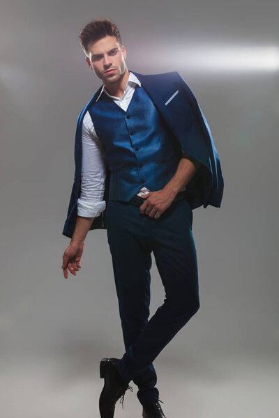 attractive young man wearing blue suit stands with legs crossed on light grey background, full body picture