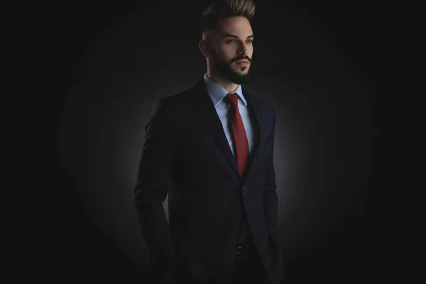 curious businessman in navy suit looks to side while standing on black background