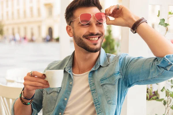 Nieuwsgierige Lachende Casual Man Vaststelling Zonnebril Drinkt Koffie Terwijl Zittend — Stockfoto