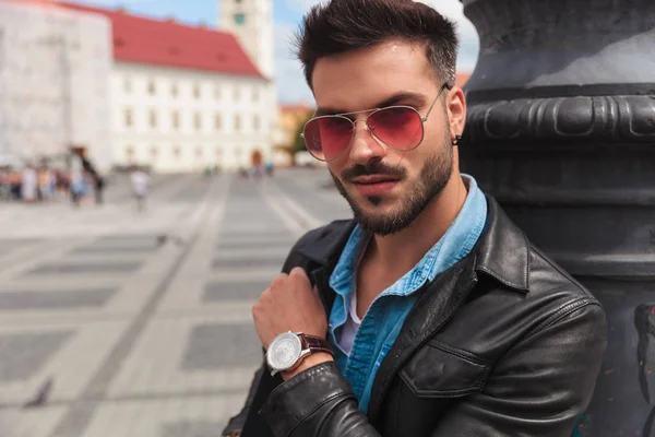 deri ceket ve kentsel arka plan üzerinde duran ve aydınlatma direği karşı yaslanmış güneş gözlüğü gülümseyen casual adam portresi