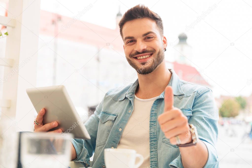 attractive casual man sitting at coffee table holds tablet and makes ok sign, portrait picture