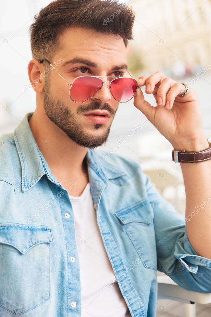 surprised seated casual man fixing red sunglasses looks to side on urban background