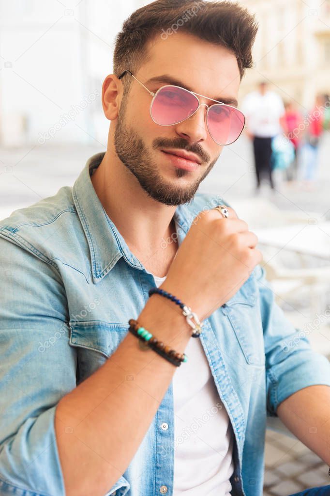 portrait of attractive casual man wearing red sunglasses sitting at table in the city smiling