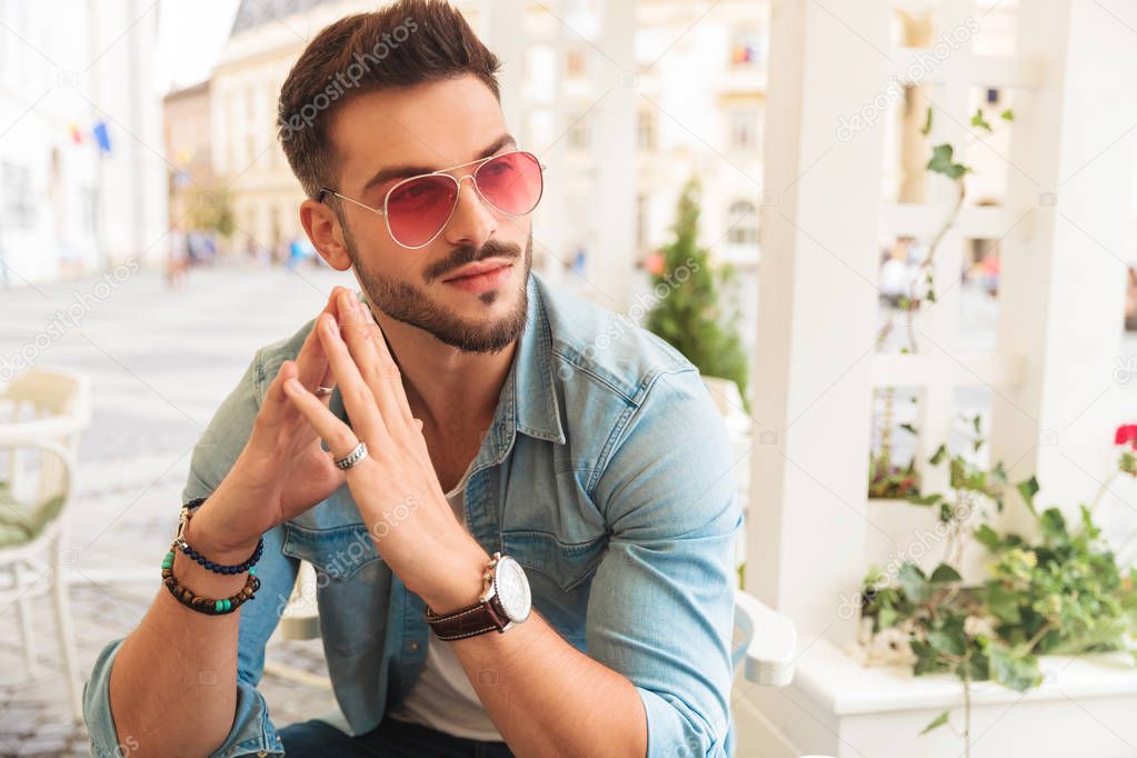 curious casual man wearing red sunglasses holding palms together while sitting at table in the city looks to side, portrait picture