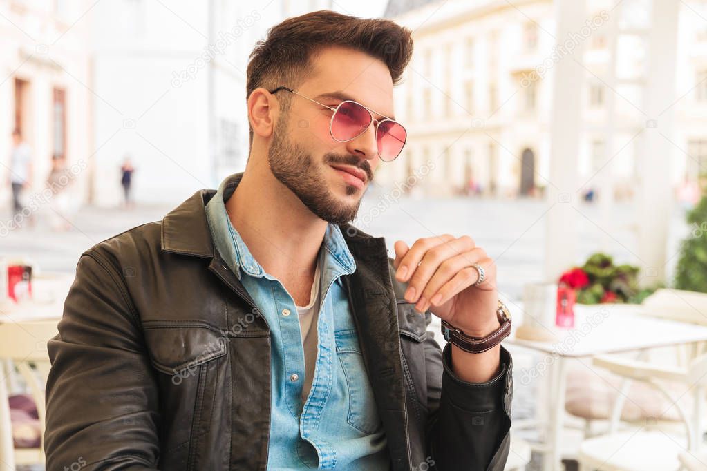 portrait of curious and smiling fashion man wearing red sunglasses and leather jacket sitting in city center at a table and looking to side