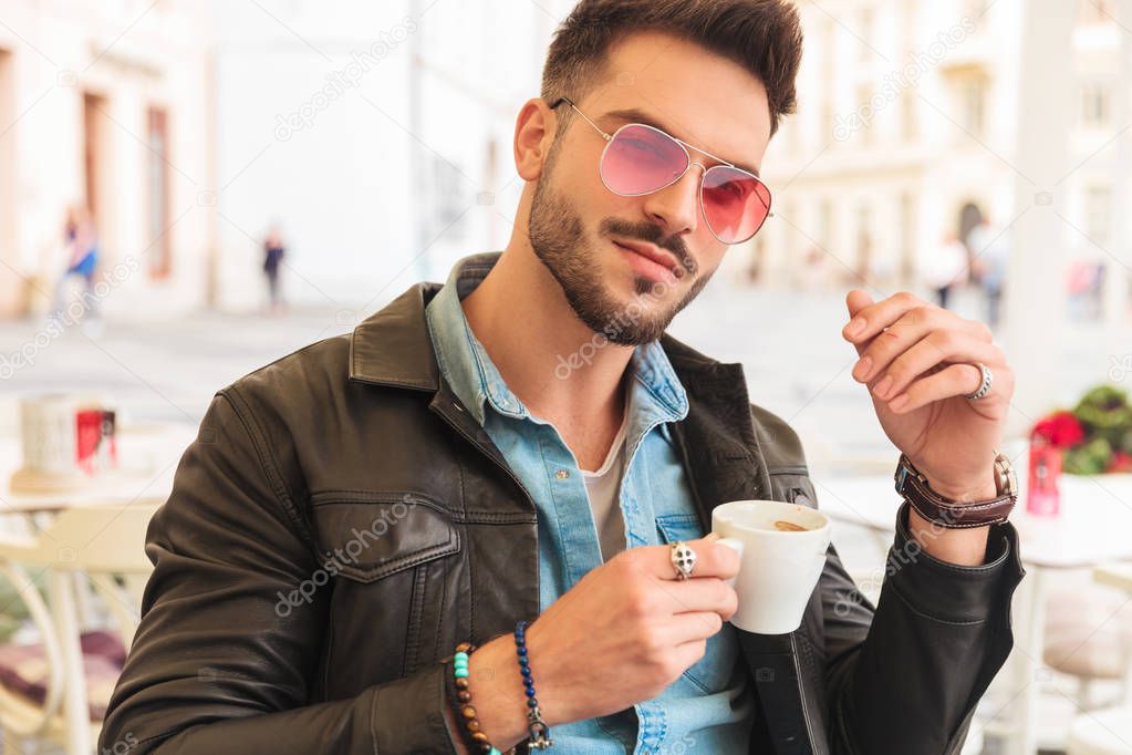 portrait of attractive casual man in leather jacket drinking coffee while sitting at a table on urban background