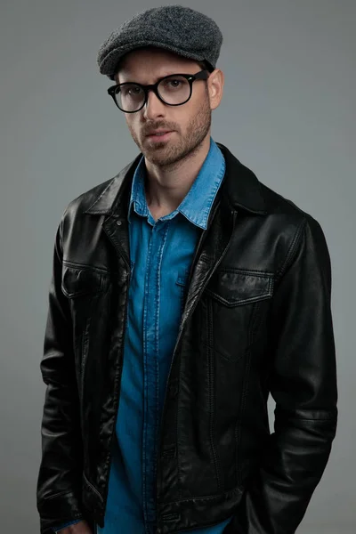 portrait of relaxed fashion man wearing leather jacket and flat cap standing on light grey background with hands in pockets