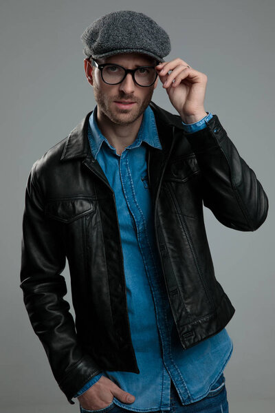 portrait of relaxed fashion man wearing leather jacket fixing eyeglasses while standing on light grey background with hand in pocket