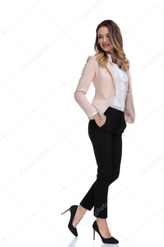 sexy smart casual woman wearing pink suit and high heels poses with feet folded and looks down to side while standing on white background, full length picture
