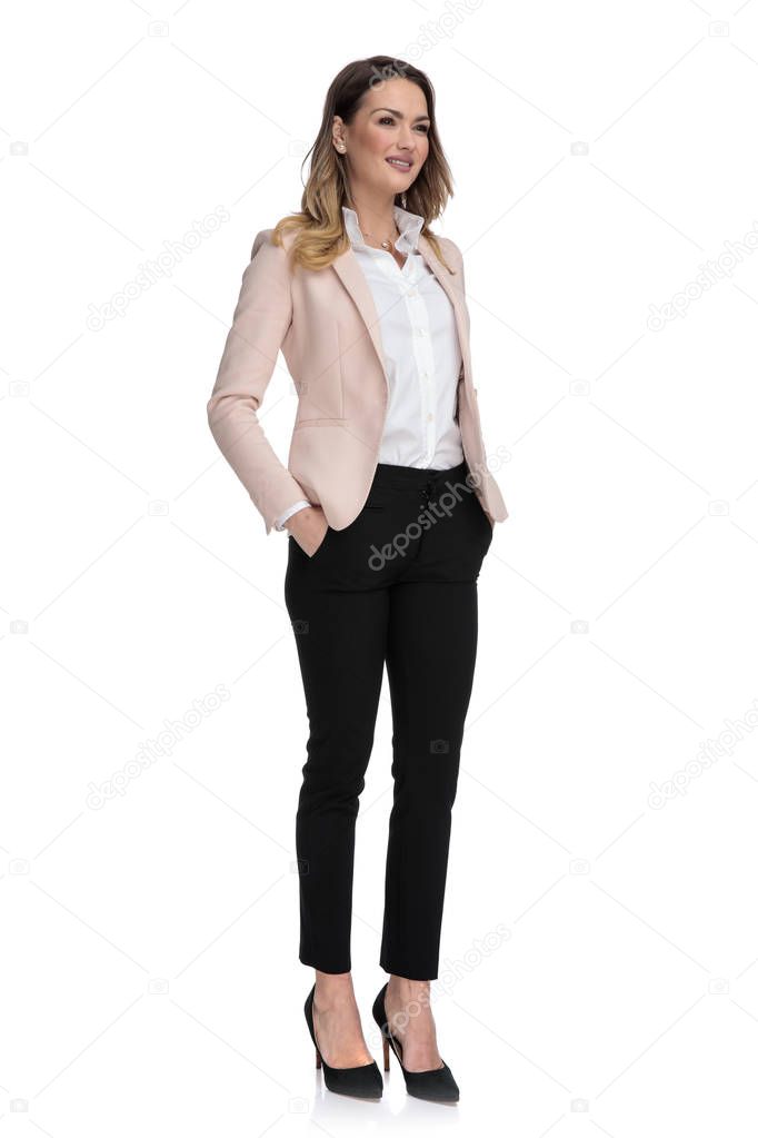 relaxed businesswoman wearing high heels and pants looks to side while standing on white background, full body picture