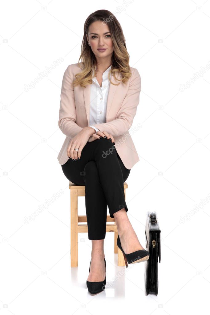 attractive businesswoman wearing pink suit sitting on wooden chair for interview with briefcase near chair on white background