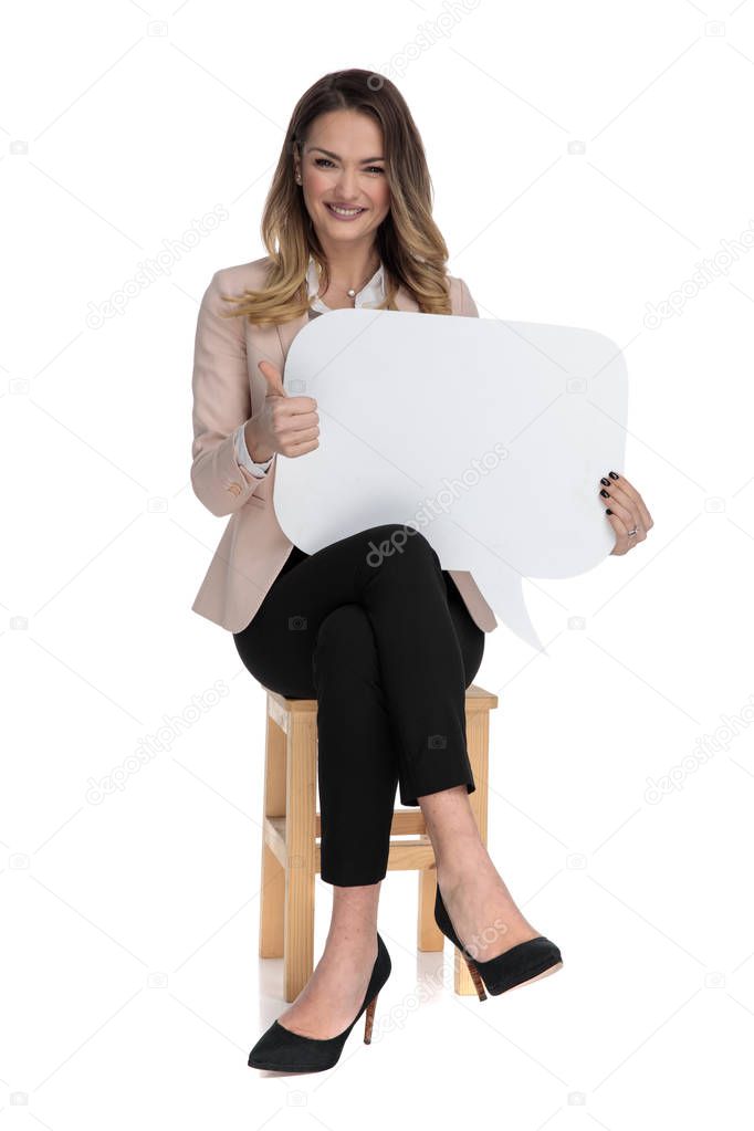 happy smart casual woman with speech bubble makes ok sign while sitting on wooden chair on white background