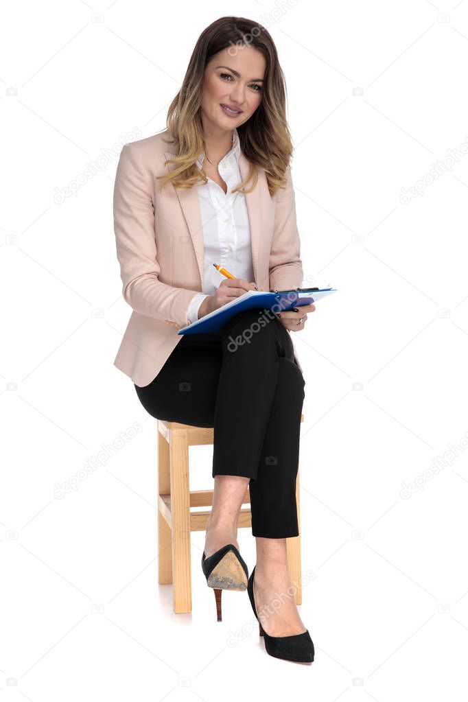 attractive businesswoman sits on wooden chair on white background and writes on blue clipboard, full body picture