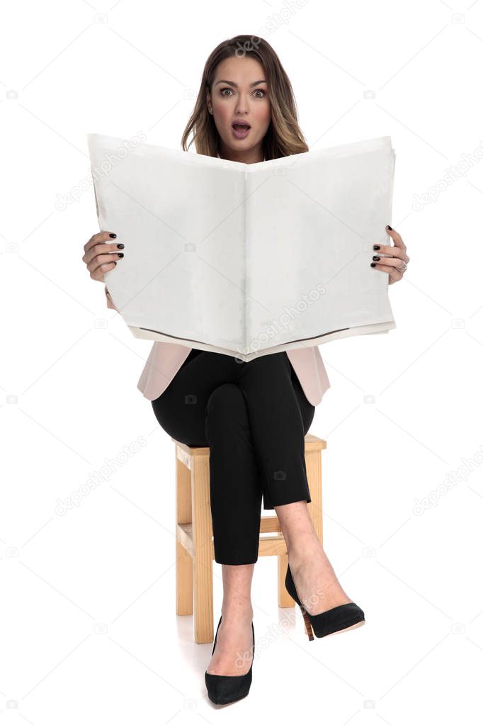 shocked businesswoman sits on wooden stool while holding newspaper on white background, full length picture