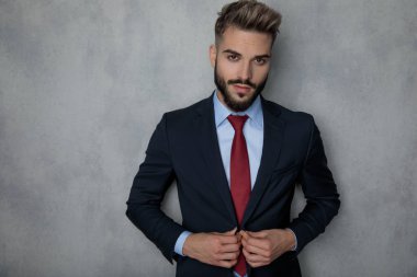 Ceketini duran gri studio duvara kadar düğmelerini gülümseyen genç moda işadamı