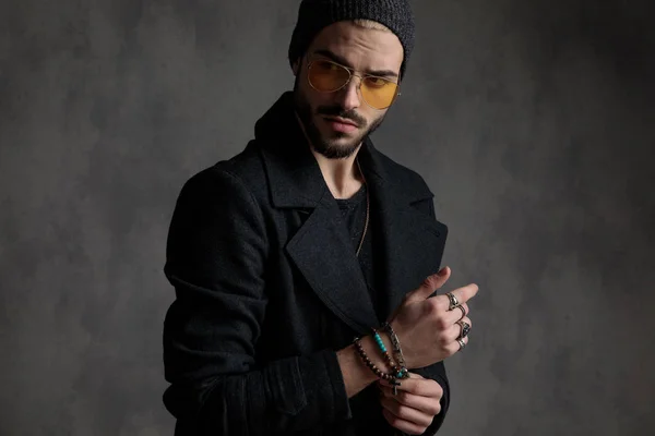Interesting man arranging his bracelets while looking at a side through a pair of sunglasses on a dark grey background