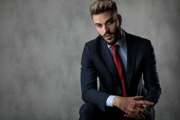 cool young businessman holding elbow on knee and palms together looks at the camera on grey background