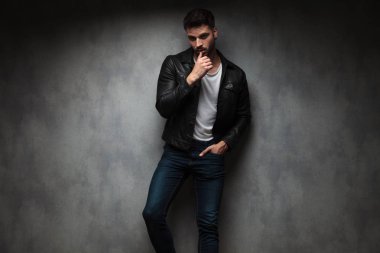 Dudaklarını parmağınızla dokunmak ve gri duvara karşı ayakta deri ceketli sesual moda adamı