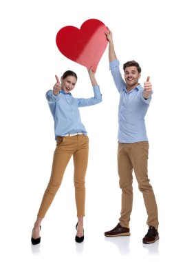Aşk yolunda! Genç mutlu çift büyük bir kırmızı kalp yukarıda başlarını tutan ve ok başparmak telafi beyaz arka plan üzerinde oturum açın