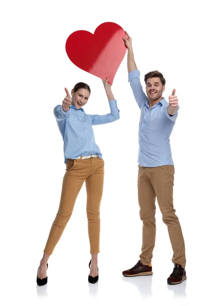 若い幸せなカップル彼らの頭の上の大きな赤いハートを押し 親指をでっちあげている白い背景の署名します — ストック写真