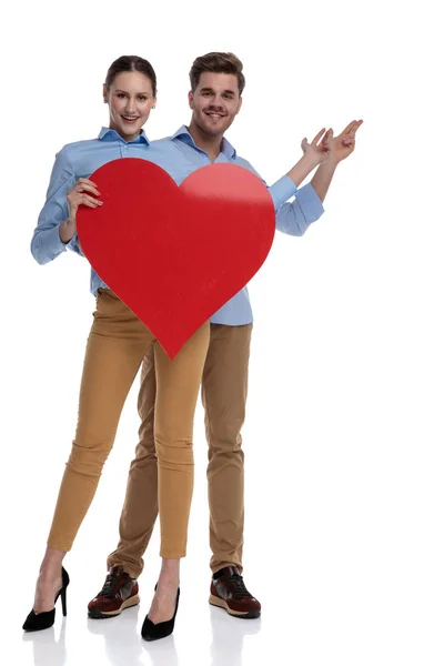 大きな赤い心を持って幸せなカップルは白地ポインティング指で何かを提示します — ストック写真