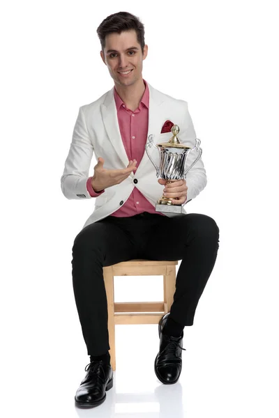 Sorrindo homem elegante sentado e apresentando seu copo de troféu — Fotografia de Stock