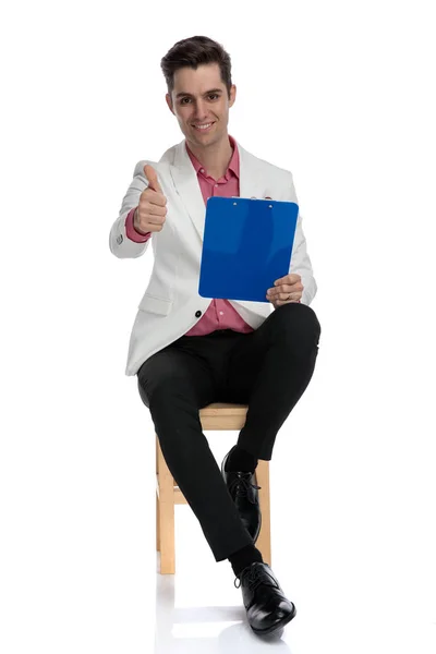 Hombre de negocios sentado en la silla sostiene un bloc de notas y hace ok —  Fotos de Stock