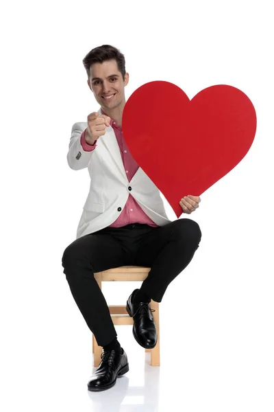 Сидячий щасливий елегантний чоловік тримає червоне серце і вказує пальцем — стокове фото
