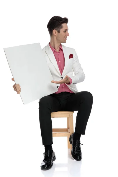 Joven hombre de negocios sentado y presentando un tablero en blanco al lado — Foto de Stock