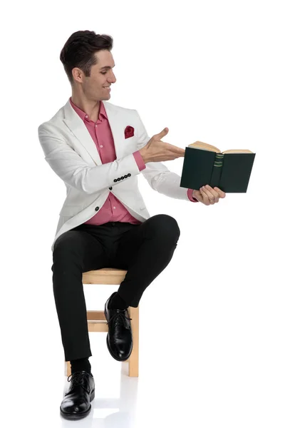 Улыбающийся молодой человек, сидя, показывает книгу в сторону — стоковое фото