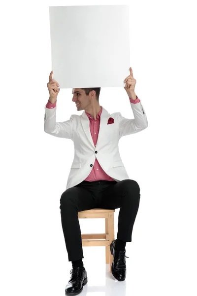 Hombre de negocios sentado sostiene anuncio por encima de la cabeza y mira a un lado —  Fotos de Stock