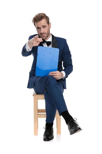 Sentado moda homem apontando dedo para câmera enquanto segurando clipb — Fotografia de Stock