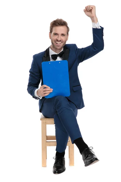 Sentado homem elegante está comemorando a vitória enquanto segurando um clipbo — Fotografia de Stock