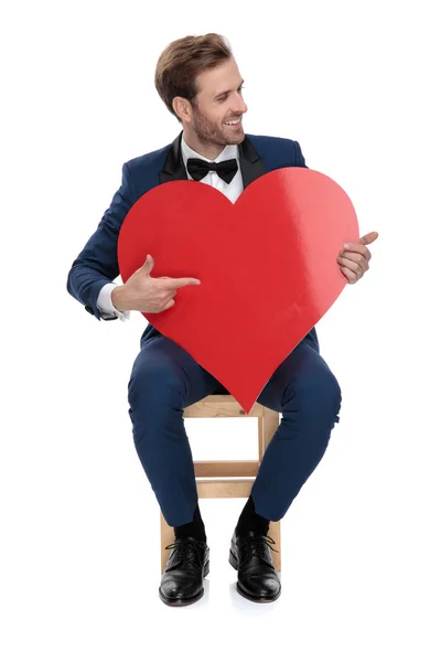 Cavalheiro sentado apontando o dedo para o coração vermelho enquanto olha para longe — Fotografia de Stock