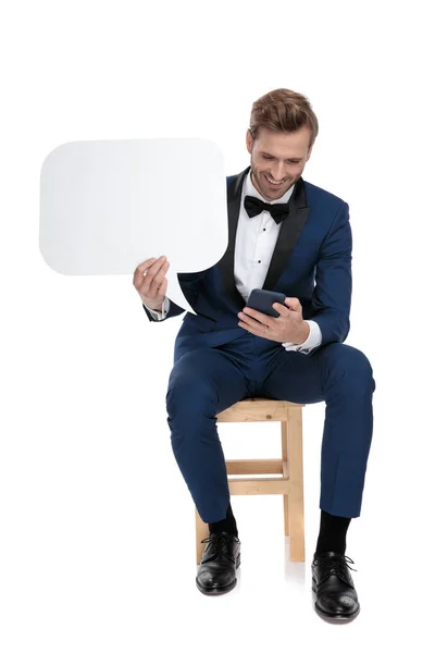 Seated attractive man texting while holding a blank speech bubbl — Stock Photo, Image