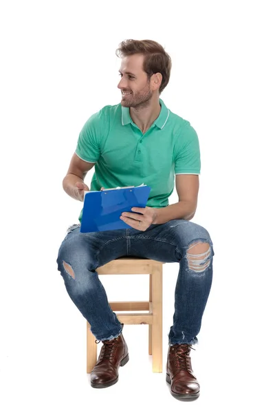 Sentado atractivo hombre en polo verde presentando un portapapeles — Foto de Stock