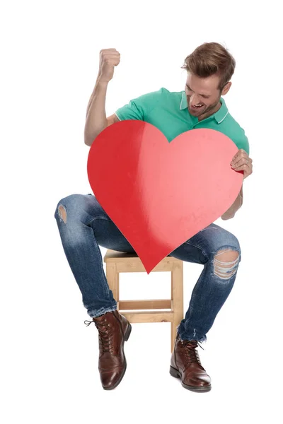 Sedící šťastný muž oslavuje vítězství rudým srdcem v rukou — Stock fotografie