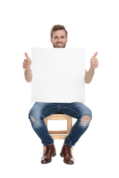 Sentado modelo homem mostra ok gesto e detém outdoor em branco — Fotografia de Stock
