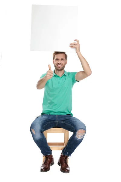 Seated sexy man shows ok gesture and holds paper board — стоковое фото