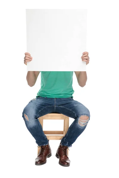 Sentado homem moderno cobre seu rosto atrás de placa de papel em branco — Fotografia de Stock