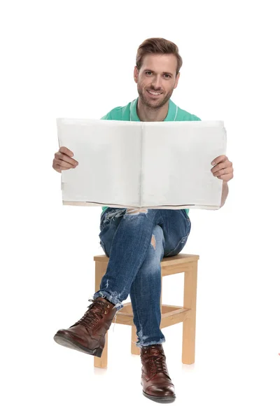 Sitzender junger Mann liest Zeitung mit gekreuzten Beinen — Stockfoto