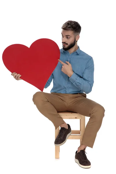 Sentado curioso hombre señalando su dedo a un corazón rojo — Foto de Stock