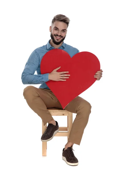 Sedící student v džínských košili má ruku na červeném srdci — Stock fotografie