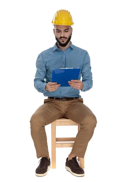 Seated focused engineer with yellow safety helmet reading from c — Stock Photo, Image