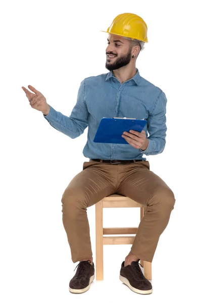 Attractive engineer sitting and greeting with a clipboard in han — Stock Photo, Image